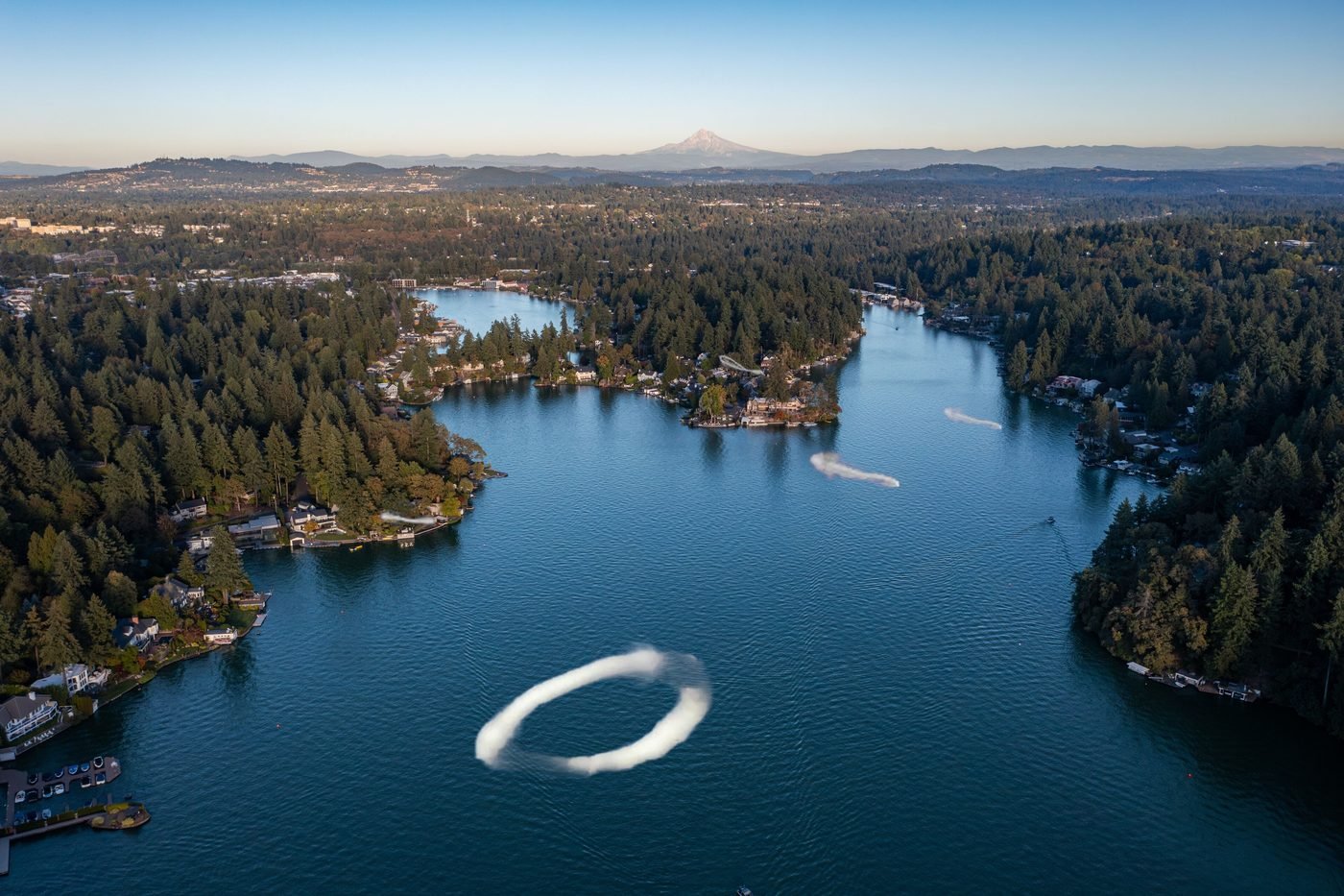 Aerial view of Lake Oswego at dawn, several smoke rings drifting low over the water toward the shoreline with Mount Hood on the horizon