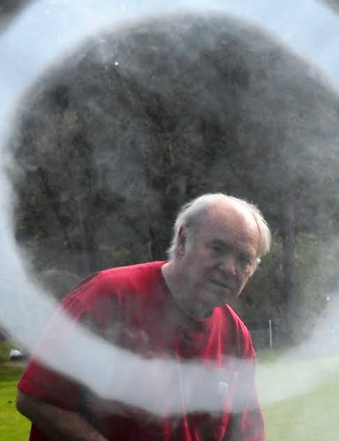A large smoke ring drifting toward the camera, with a white-haired man in a red shirt watching from behind it