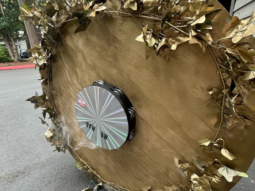Prototype VII: a large gold-toned circular drum wreathed in ivy garland, with a mirrored tambourine mounted at its center reading MR MAN, faint smoke drifting from the base